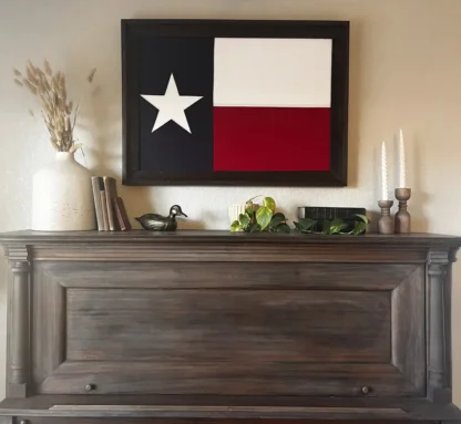 Framed Texas flag hanging on a beige wall above a vintage wooden piano, decorated with rustic accents including books, candles, a ceramic vase with dried plants, and a small potted plant.